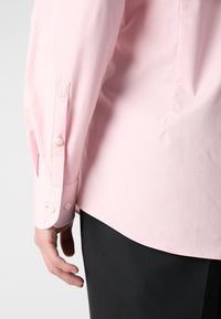 Personne portant une chemise rose clair à manches longues avec des poignets boutonnés et un pantalon noir, vue de dos et de la taille vers le bas.