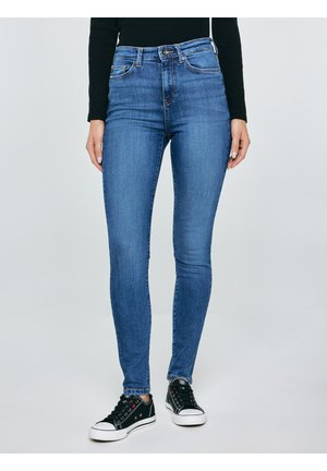 Žena v modrých úzkých džínách s vysokým pasem a černých nízkých teniskách stojí před jednobarevným světle šedým pozadím.