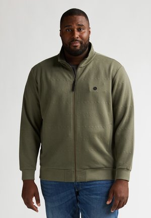 Olijfgroene jas met ritssluiting, voorzien van een gestructureerd patroon, geribbelde manchetten en kraag. Draagt over een donker shirt. Heeft een klein logo op de borst.