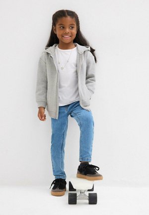 Bambino con una felpa grigia con zip sopra una maglietta bianca, jeans blu e sneaker nere con suola beige, in piedi su uno skateboard.