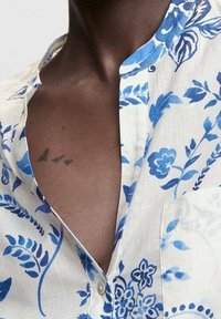 Chemise en lin blanc avec un motif floral bleu, col mandarin et un détail de bouton. Tatouage visible d'oiseaux près de la clavicule.