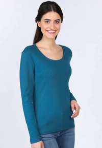 Langarm-Tee in Teal mit Rundhalsausschnitt, strukturiertem Punktmuster und lässiger Passform. Kombiniert mit blauen Jeans.