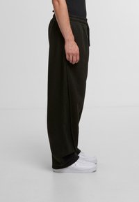 Dark green sweatpants with elastic waistband, side pockets, and a loose fit, paired with white athletic shoes. Smooth fabric texture.