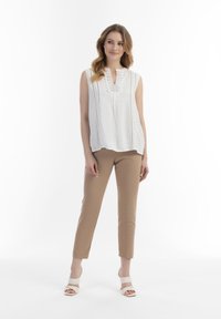 Witte mouwloze top met ingewikkelde voorzijde, gedragen met lichtbruine aansluitende broek, afgewerkt met witte sandalen met bandjes.