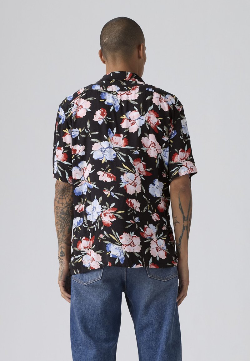 Mand med kort hår og tatoveringer iført en sort kortærmet skjorte med rød, pink, blå og hvid blomstret print og blå jeans, set bagfra.
