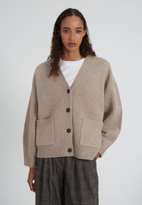 Cardigan beige en tricot avec col en V, boutons noirs et deux poches avant. Poignets et ourlet côtelés, avec une coupe décontractée.