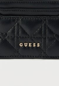 Μαύρο δερμάτινο ύφασμα με μικρές ραφές σε σχήμα καρδιάς και γράμματα από χρυσό μέταλλο που σχηματίζουν τη λέξη "GUESS" στην επιφάνεια τσάντας ή πορτοφολιού.