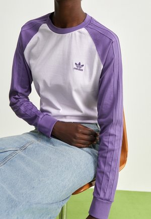 Person iført en hvid og lilla Adidas langærmet trøje og lyseblå jeans, siddende på en træstol mod en ensfarvet baggrund.