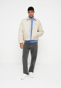 Šviesiai rudas pūkinis švarkas su užtrauktuku priekyje ir stovinčiu apykakle, derinamas su mėlynu džemperiu ir pilkais džinsais. Modelis dėvi baltus sportbačius.