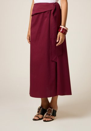 Jupe enveloppante bordeaux avec un détail de lien sur le côté, en tissu lisse. Portée avec des talons ouverts noirs mettant en valeur des accents dorés et des bracelets empilés.