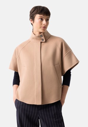 Persoon draagt een beige jas met korte mouwen, hoge kraag en knopen, een zwart shirt met lange mouwen eronder en donkere gestreepte broek, handen in de zakken.