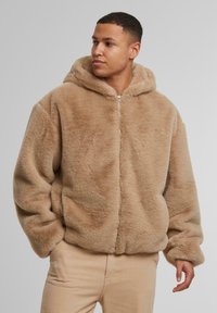 Beige Kunstfell-Hoodie mit einem Front-Reißverschluss, oversized Passform und großer Kapuze. Der Stoff hat eine weiche, flauschige Textur und elastische Bündchen.