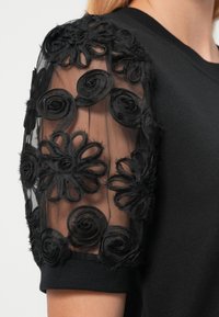 Μαύρο τοπ με κοντά διαφανή μανίκια στολισμένα με υφασμάτινα λουλούδια και κέντημα. Το σώμα έχει υφή σταθερού μαύρου πλεκτού.