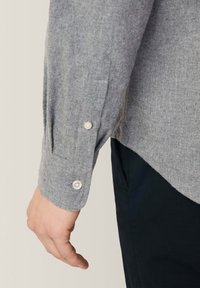 Grijze, gestructureerde, langelijf shirt met knoopmansetten, voorzien van contrasterende lichte knopen en een iets gebogen zoom tegen donkere broeken.