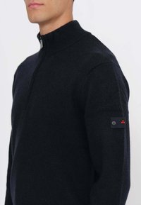 Uomo che indossa un maglione nero con mezza cerniera, colletto a coste e una toppa con bottone e tre puntini rossi sul braccio sinistro superiore.