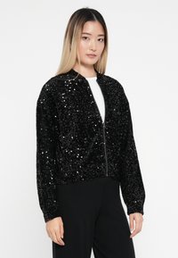 Czarna kurtka bombowa pokryta cekinami, z zamkiem z przodu, długimi rękawami i miękką fakturą. Odpowiednia na różne okazje.