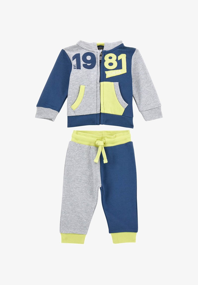 Pilkas ir tamsiai mėlynas treniruoklių komplektas su žaliais akcentais. Yra džemperis su užtrauktuku, gobtuvu, kišenėmis ir spaudiniu "1981". Kelnės turi raištelį ir siaurėjantį siluetą.