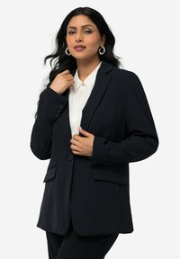 Blazer marine avec un design structuré, des revers crantés, deux boutons et des poches latérales. Porté sur une chemise blanc à col. Texture lisse.