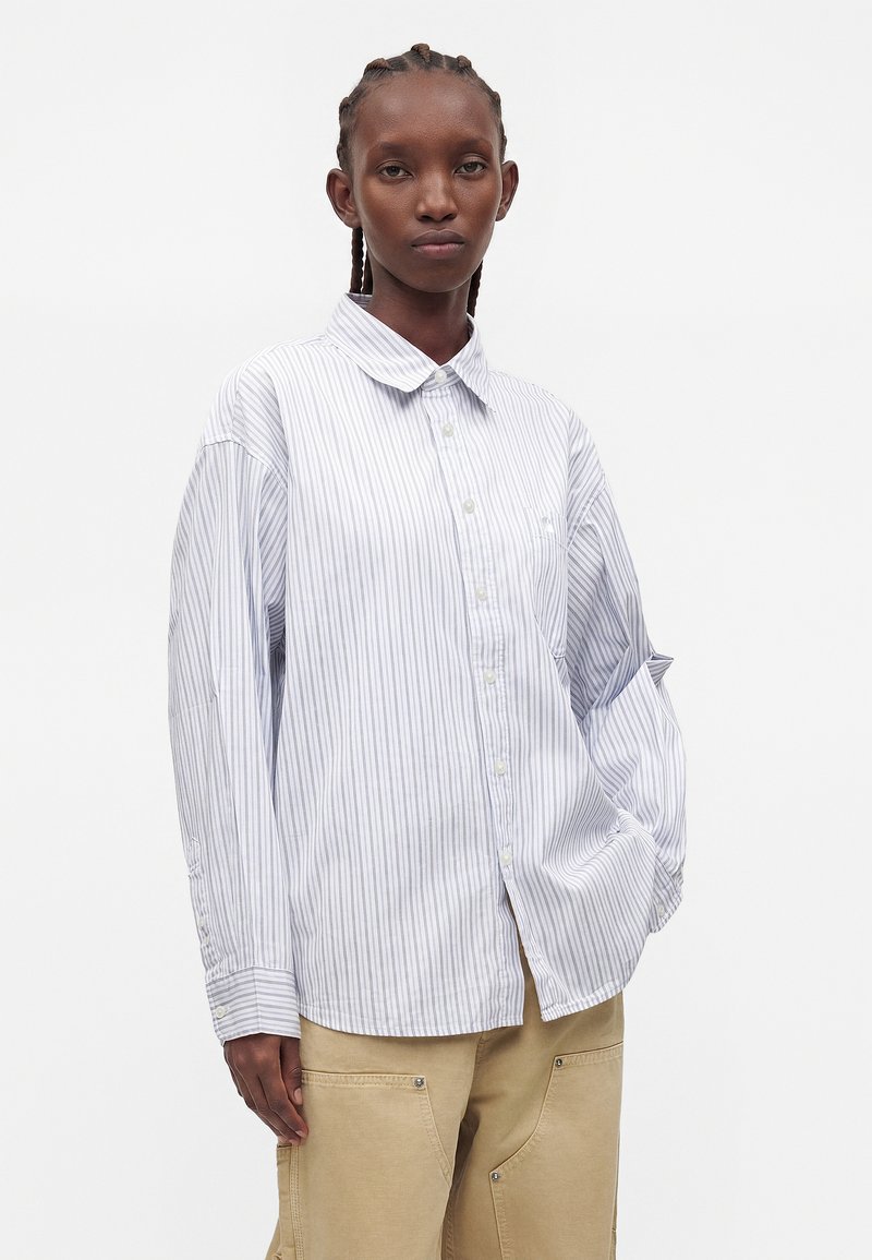 Donna con i capelli intrecciati che indossa una camicia bianca e blu a righe larga con bottoni e pantaloni beige, in piedi davanti a uno sfondo semplice.