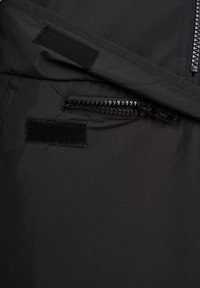 Tissu noir avec une poche à fermeture éclair partiellement ouverte, deux bandes velcro noires, et un rabat plié au-dessus de la poche.