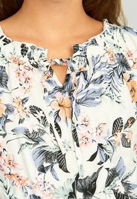 Blouse fleurie en tissu doux avec un fond blanc orné de fleurs roses et bleues, encolure élastique et détail à nouer. Découpe en V sous le col.