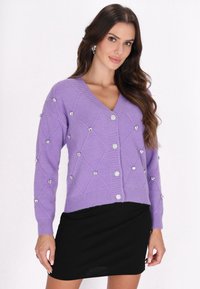 Lila gestrickte Strickjacke mit V-Ausschnitt, Rautenmuster, verziert mit blumenförmigen Knöpfen, kombiniert mit einem schwarzen Rock.