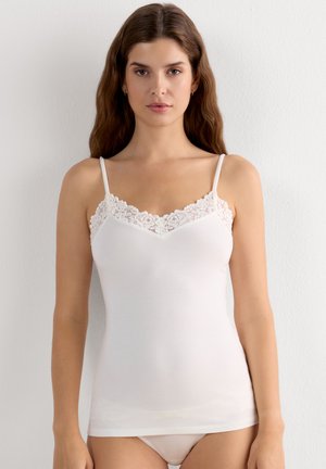 Žena s dlouhými vlasy nosící bílý top s krajkovým lemem a odpovídající bílé spodní prádlo, stojící před jednoduchou světlou zdí.