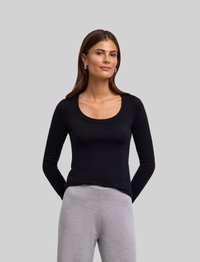 Schwarzes Langarmshirt mit einem Rundhalsausschnitt, aus weichem Material. Getragen mit hellgrauen, strukturierten Hosen hat das Modell die Hände hinter dem Rücken.
