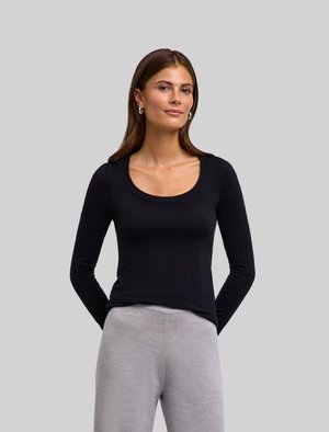 Zwarte langemouwen top met een ronde hals, gemaakt van zachte stof. Draagt een lichtgrijze, gestructureerde broek, terwijl het model haar handen achter haar rug heeft.