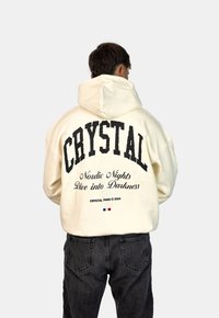 Felpa con cappuccio in bianco sporco, con grande testo nero "CRYSTAL" e testo aggiuntivo sotto. Tessuto spesso, vestibilità comoda, indossata con jeans scuri.