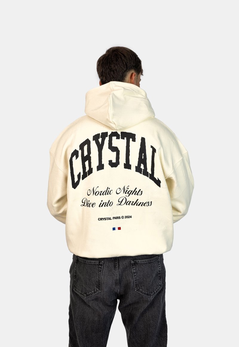 Felpa con cappuccio in bianco sporco, con grande testo nero "CRYSTAL" e testo aggiuntivo sotto. Tessuto spesso, vestibilità comoda, indossata con jeans scuri.