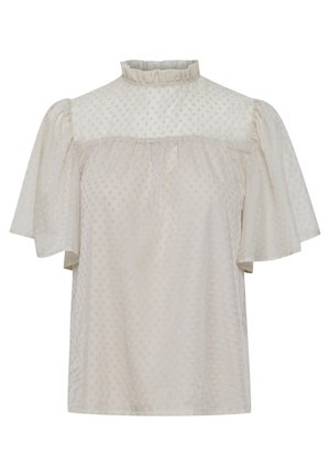 Femme portant un chemisier blanc transparent à col froufrou avec des manches papillon, un jean skinny bleu foncé et des bottines noires, debout devant un fond uni.