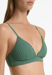 Triangle bralette od teksturiranog zelenog materijala s tankim podesivim trakama i suptilnim uzorkom. Ima nisko izrezan dekolte.