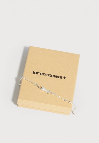 Ασημένιο βραχιόλι με αλυσίδα σε σχήμα ψαριού, τοποθετημένο πάνω σε μπεζ κουτί κοσμημάτων Loren Stewart σε λευκό φόντο.