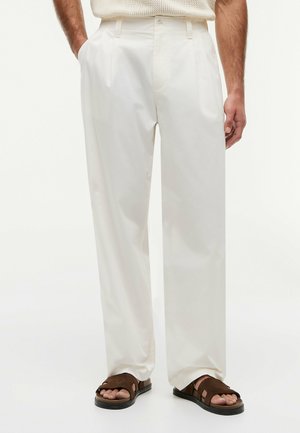 Uomo con pantaloni bianchi larghi e sandali in camoscio marrone, in piedi con le mani in tasca davanti a uno sfondo chiaro uniforme.