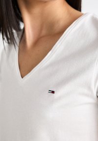 Bílý bavlněný tričko s výstřihem do V, které má na levé části hrudi malé vyšívané logo s modrými, červenými a bílými pruhy. Měkká textura.