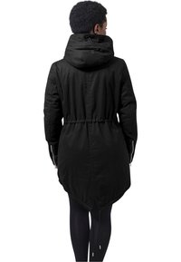 Parka noire à capuche avec une taille cintrée, des manches longues et un dos plus long. Présente des poches zippées et un ourlet arrondi. Texture lisse.