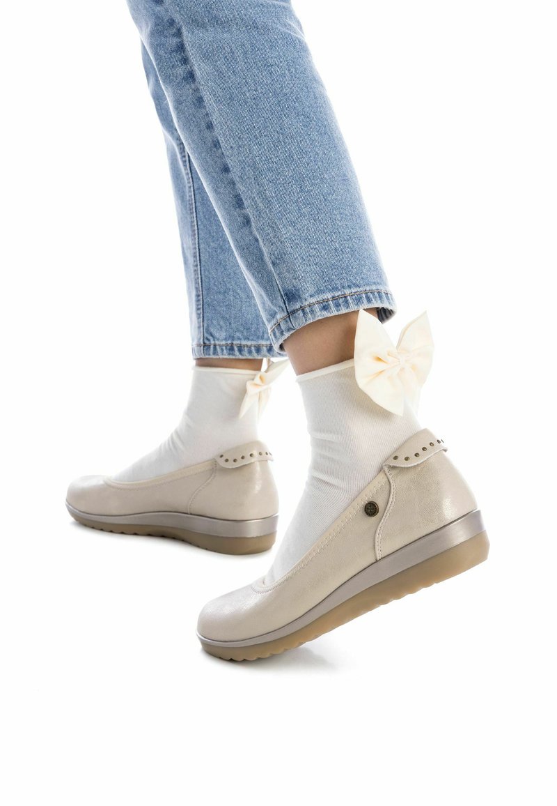 Zapatos de cuero beige con punta redondeada, superficie texturizada y tachuelas decorativas en el tacón, combinados con calcetines blancos que presentan detalles de lazo.