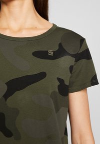 Olivově zelené tričko s černým maskáčovým vzorem, kulatým výstřihem a krátkými rukávy. Obsahuje malé zlaté logo "RAW" na hrudi.