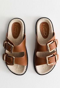 Sandalias de imitación de cuero marrón con dos hebillas doradas ajustables, diseño de punta abierta y plantilla texturizada beige sobre una suela de goma negra.