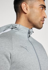 Grå zip-up hoodie med tekstureret stof, der har en hvid streg langs skulderen og et diskret Puma-logo på brystet. Justerbar hætte.