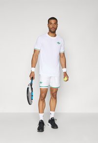 T-shirt de sport blanche avec des accents verts, short blanc assorti et baskets noires. Tenant une raquette de tennis et une balle de tennis jaune.