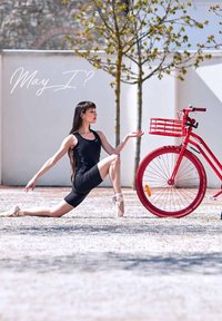 Zwart sportieve bodysuit, tan ballet schoen, en een felrode fiets met een voorrek. Open ruimte met verspreide kiezels en minimale bomen.