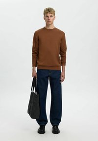 Hnědá bavlněná mikina, tmavě modré volné džíny, černé boty, drží šedou látkovou tašku s černými uchy, minimalistický design.