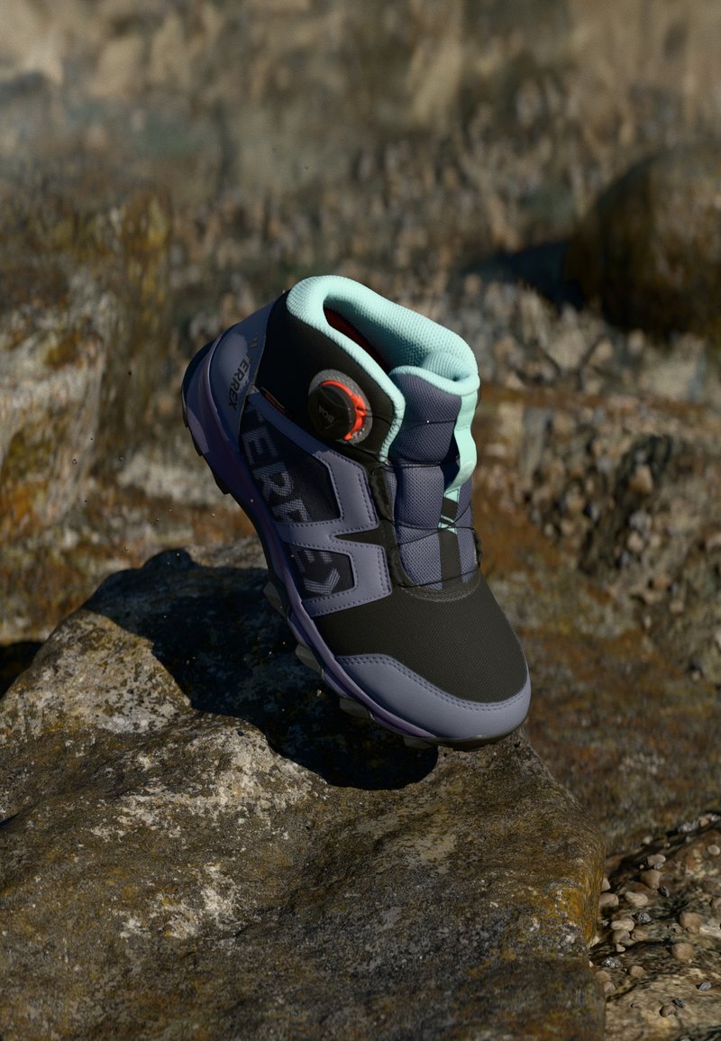 Black and gray hiking boot with turquoise accents. Features textured material, rubber sole, and a quick-lace toggle system. Positioned on a rocky surface.