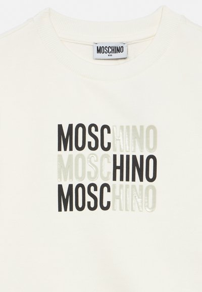 Κρεμ πουκάμισο με την επιγραφή "MOSCHINO" επαναλαμβανόμενη τρεις φορές σε μαύρα και λευκά μπλοκ γράμματα, η ετικέτα γράφει "MOSCHINO KID" στον γιακά.