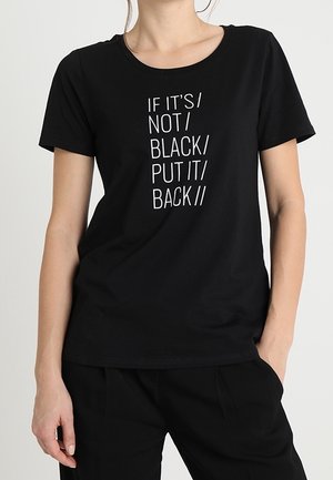 Persona que lleva una camiseta negra con texto blanco que dice "IF IT'S / NOT / BLACK / PUT IT / BACK" y pantalones negros, con las manos en los bolsillos.