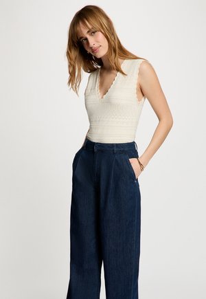 Jonge vrouw met schouderlang blond haar die een mouwloze witte textuurtop draagt en hooggetailleerde wijde donkere jeans.