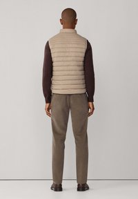 Chaleco acolchado beige con diseño de acolchado, llevado sobre un suéter oscuro. Pantalones ajustados marrones y zapatos marrones completan el atuendo, visto desde la espalda.