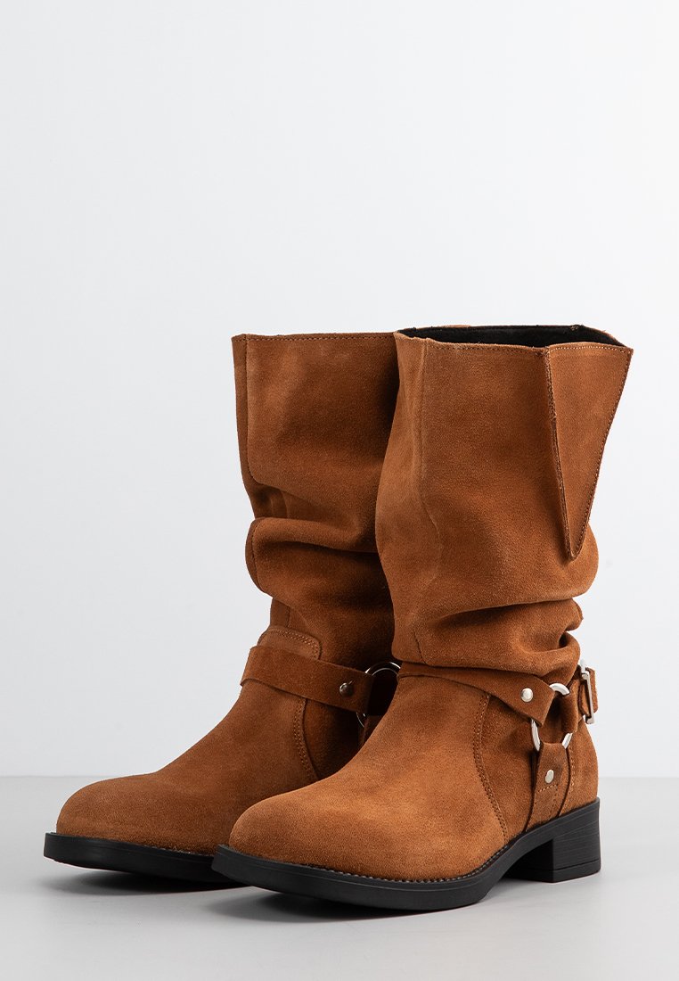 Braune Wildleder-Ankle Boots mit einem lässigen Design, ausgestattet mit einem dekorativen Riemen und silbernen Hardware-Akzenten, auf einem niedrigen schwarzen Absatz.